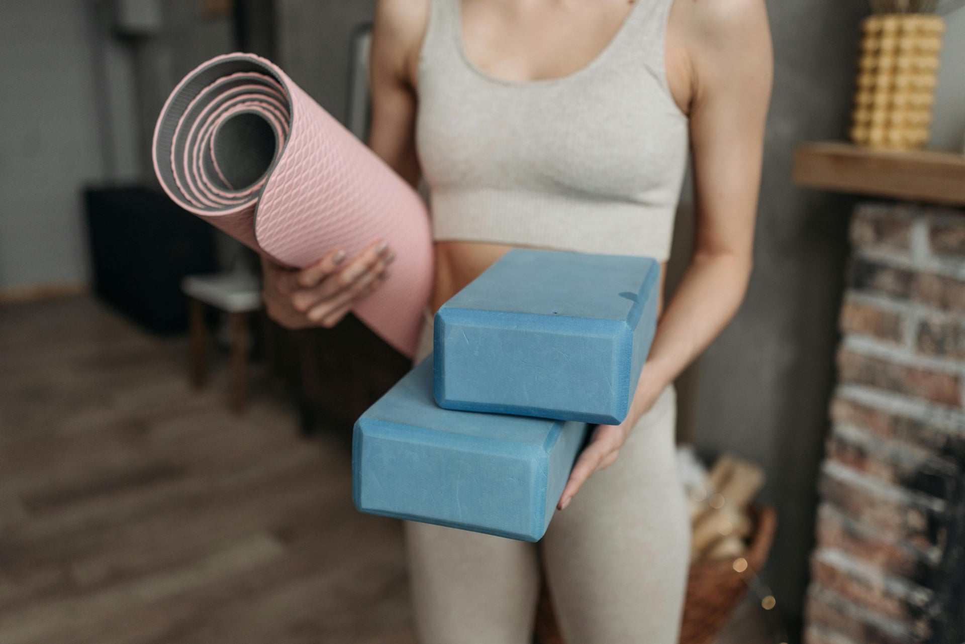 A young woman holding a pink yoga mat and blue yoga blocks in a cozy indoor setting, ready to start a yoga practice.