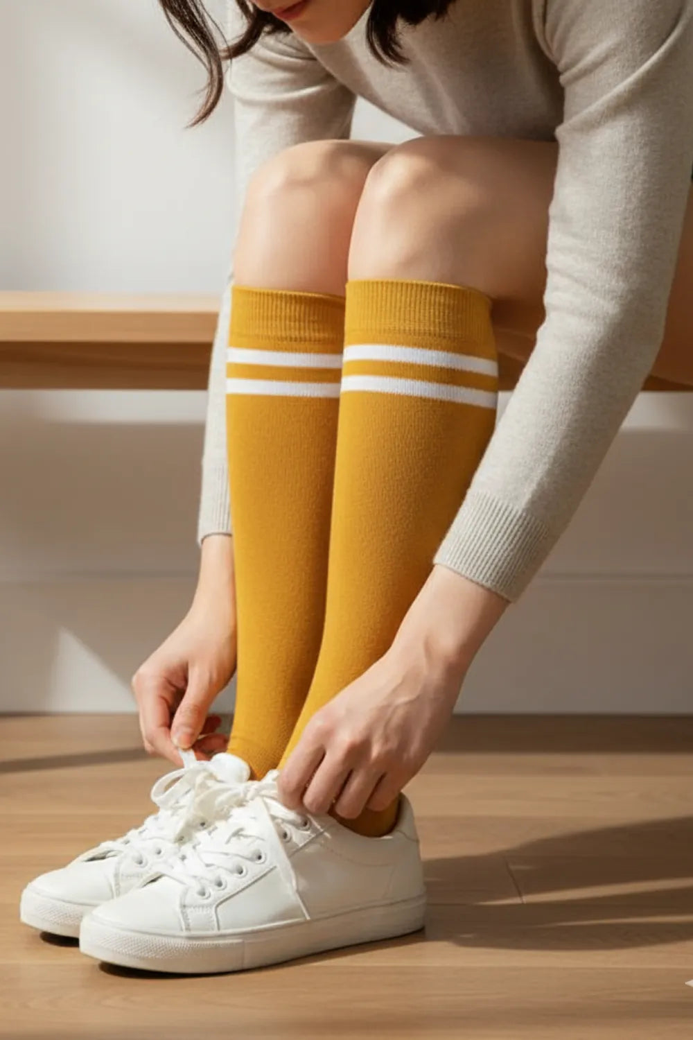 Mustard yellow striped knee-high socks while tying white sneakers on a wooden bench.