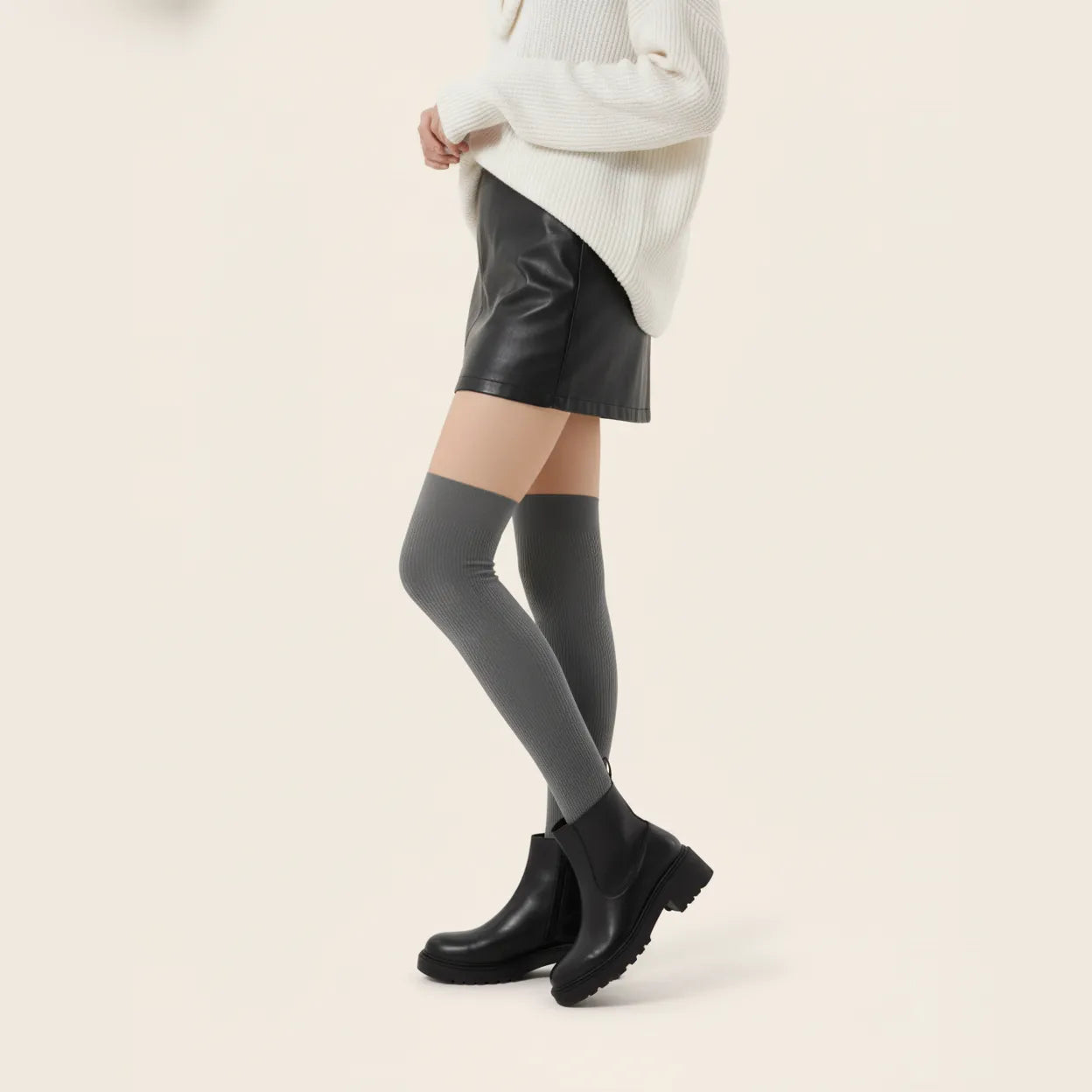 Woman styling dark grey ribbed over-the-knee socks with a cream knit sweater, black leather-look mini skirt and ankle boots.