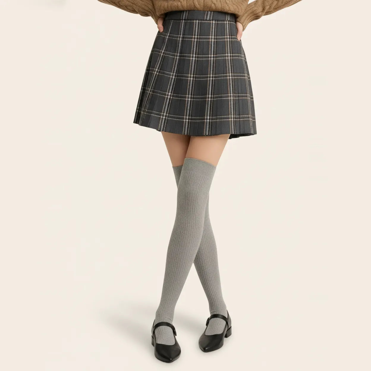 Woman styling grey ribbed over-the-knee socks with a grey plaid pleated skirt, brown knit sweater and black Mary Jane shoes.