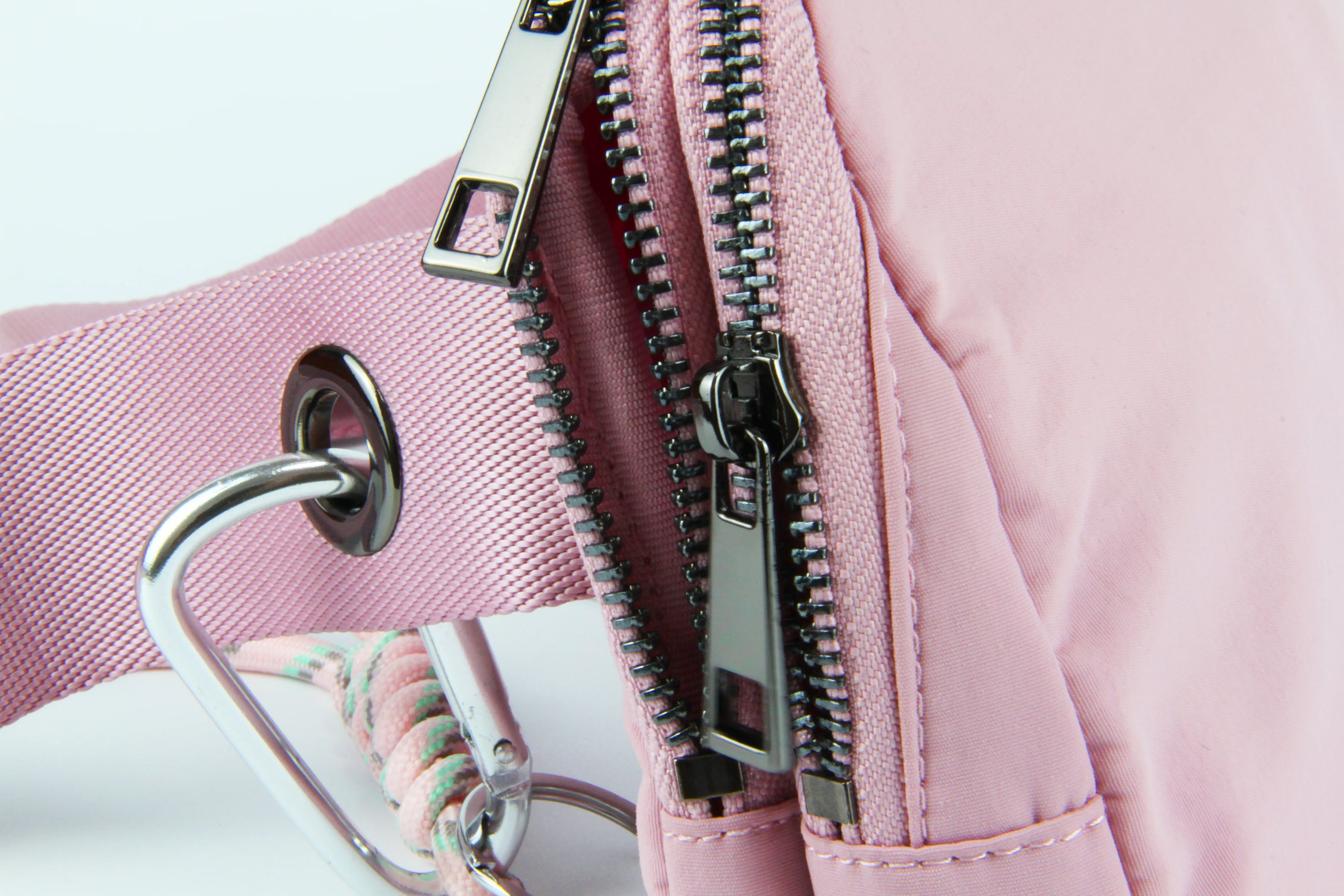 Close-up of a blush-pink water-resistant nylon belt bag showing dual metal zippers, reinforced grommet, and a detachable carabiner with braided cord.