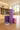 SportiveLives yoga setup in a sunlit studio: a lavender textured foam roller stands beside a deep-violet yoga block and a dusty-rose pink yoga block, with a lilac-and-white numbered stretch strap on a wooden floor; shelving with rolled mats in the background.