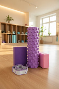 SportiveLives yoga setup in a sunlit studio: a lavender textured foam roller stands beside a deep-violet yoga block and a dusty-rose pink yoga block, with a lilac-and-white numbered stretch strap on a wooden floor; shelving with rolled mats in the background.